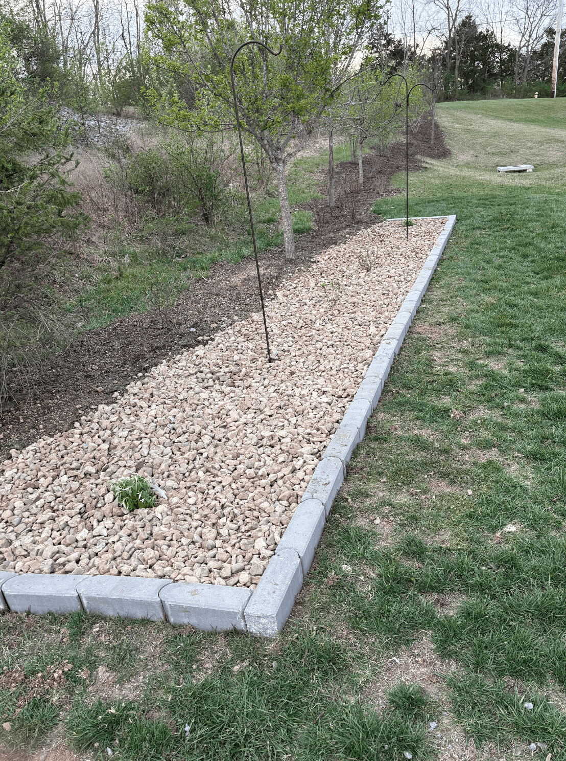 Long rectangular decorative rock bed with block edging running along a tree line